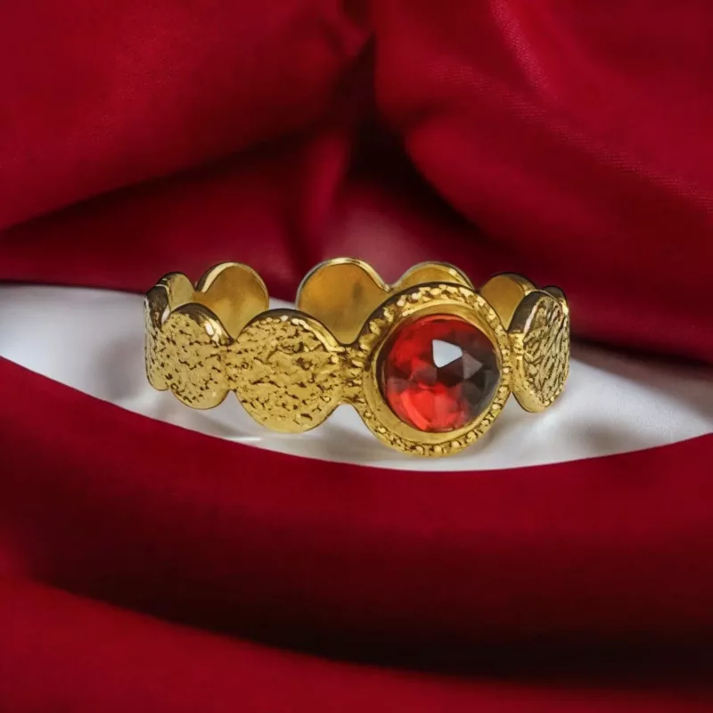 Une bague dorée ornée d’une pierre naturelle rouge centrale, entourée de motifs texturés, présentée sur un fond en tissu rouge et blanc.