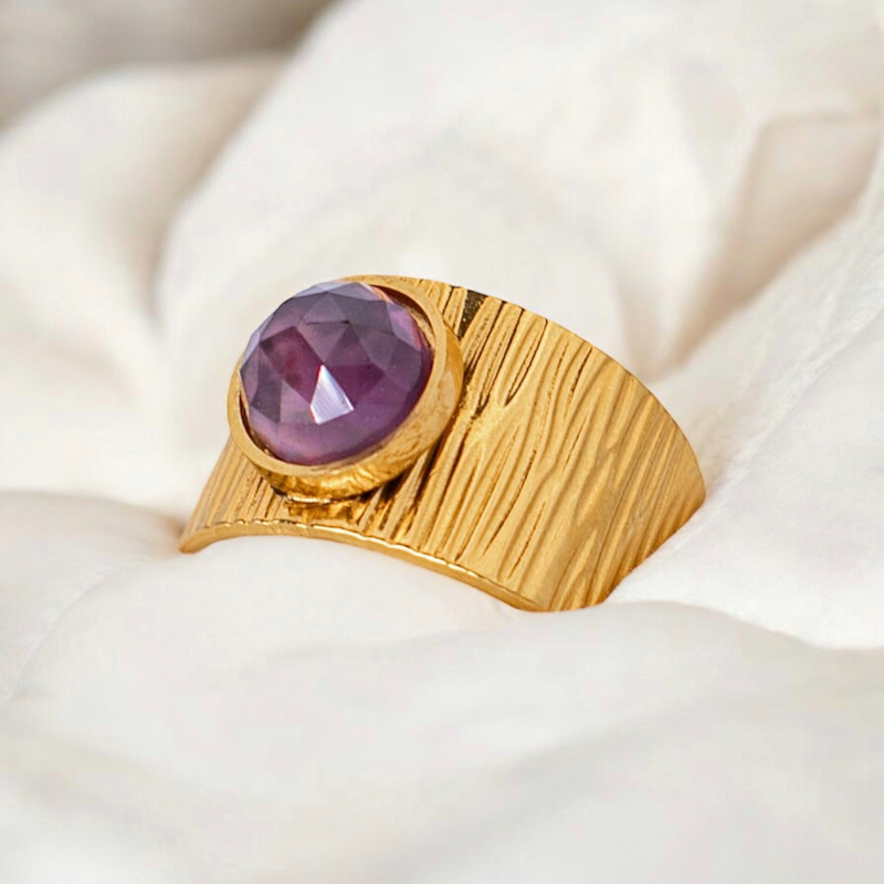 Une bague en or texturé ornée d'une améthyste violet, posée sur un fond doux et neutre.