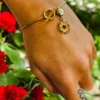 Bracelet doré délicat avec un charm en forme d’étoile à huit branches et une pierre rouge, porté sur un poignet devant un fond de fleurs rouges.