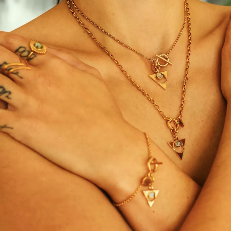 Collection chakras Vishuddha : Gros plan sur un modèle portant des bijoux dorés raffinés, comprenant des colliers et un bracelet avec des pendentifs triangulaires ornés de topaze bleue, accompagnés de bagues assorties dans un style chic et moderne.