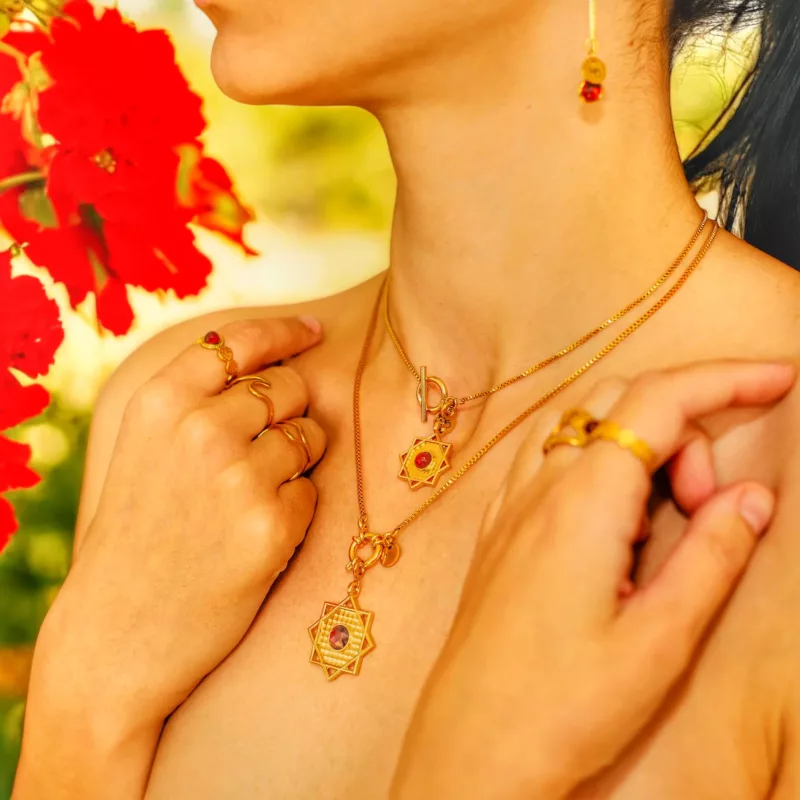 collection chakras muladhara : Femme portant des bijoux en or ornés de pierres naturelles rouges (grenat), avec un collier en étoile, des bagues et des boucles d’oreilles, devant un fond fleuri rouge.